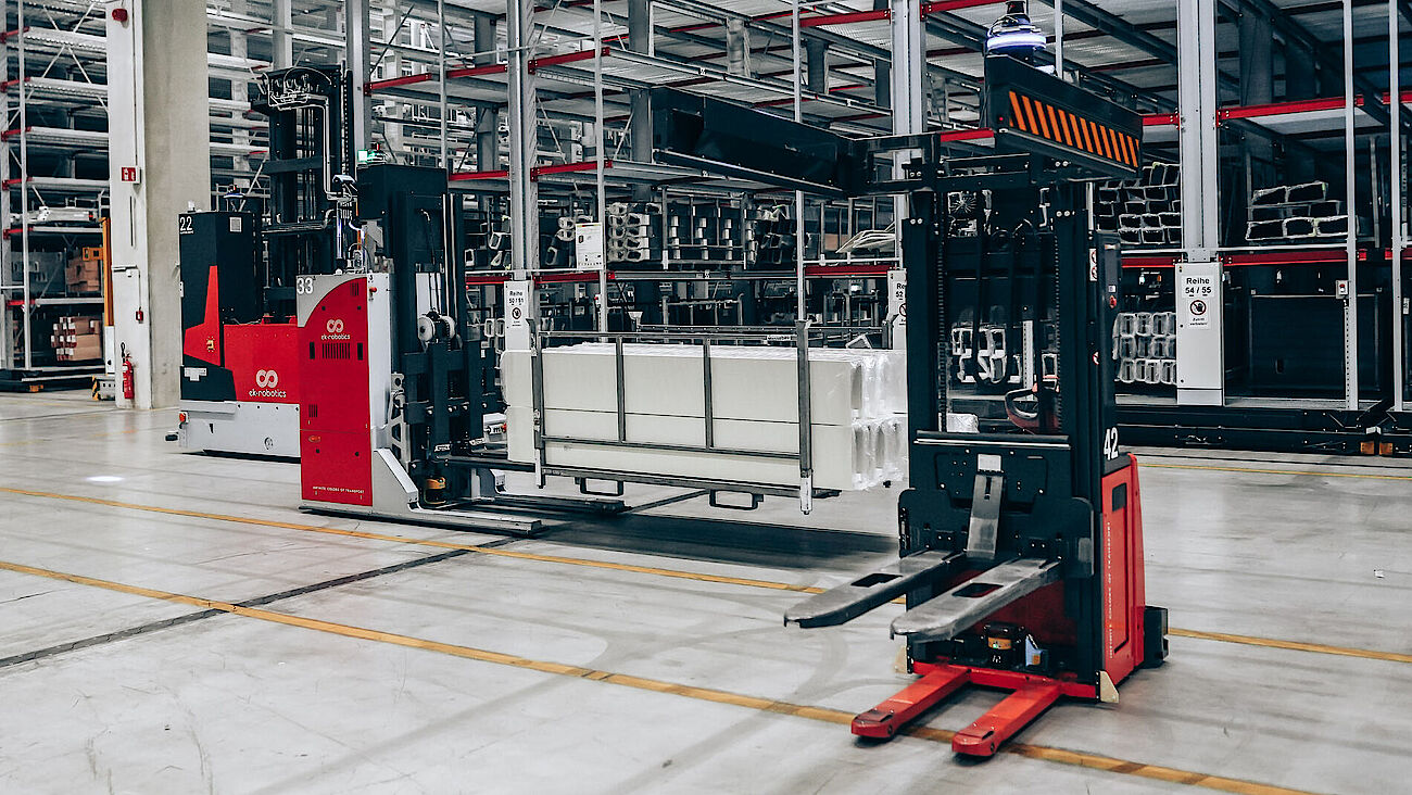 Drei rote Transportroboter von ek robotics bewegen Güter vor einer Regalanlage.